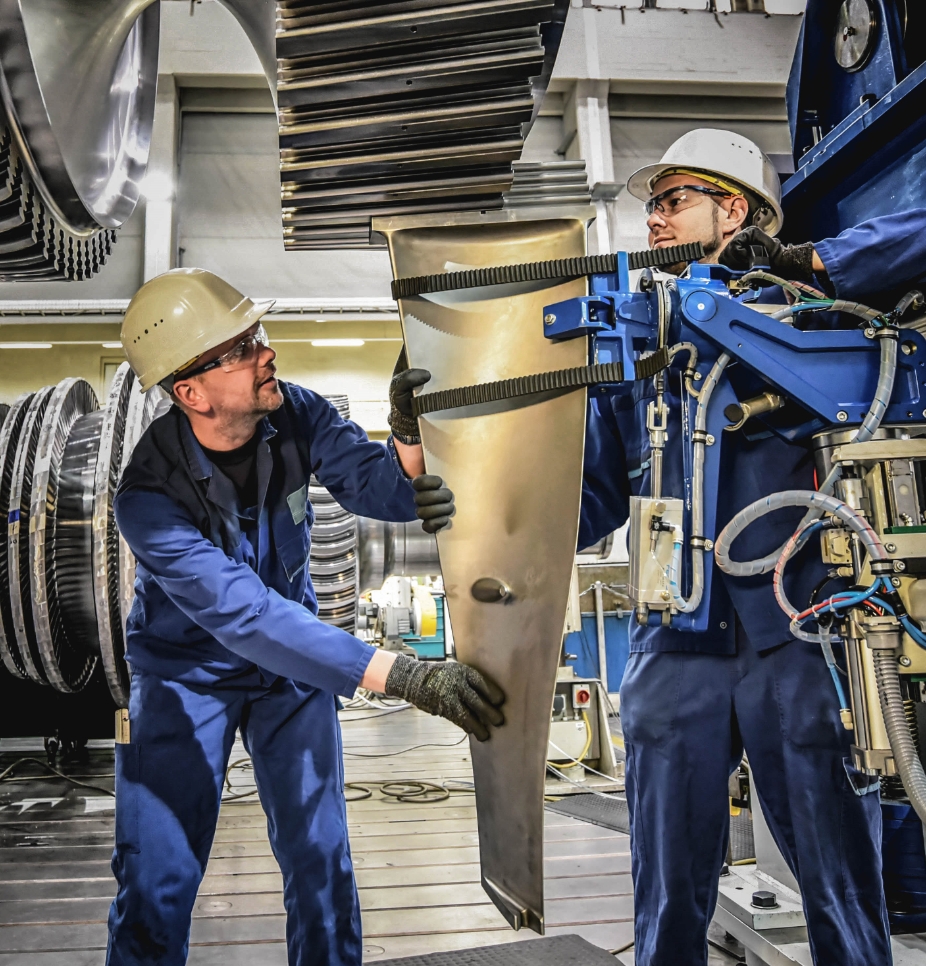 Two men in blue overalls, with white hard hats and safety googles, working in a factory for turbines