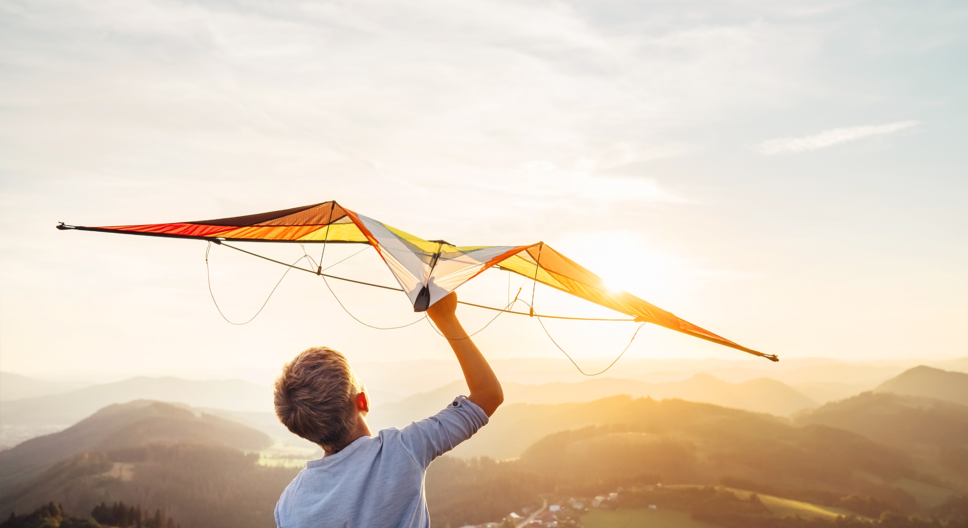 A person at the top of a hill, holding a kite and looking towards the sunset