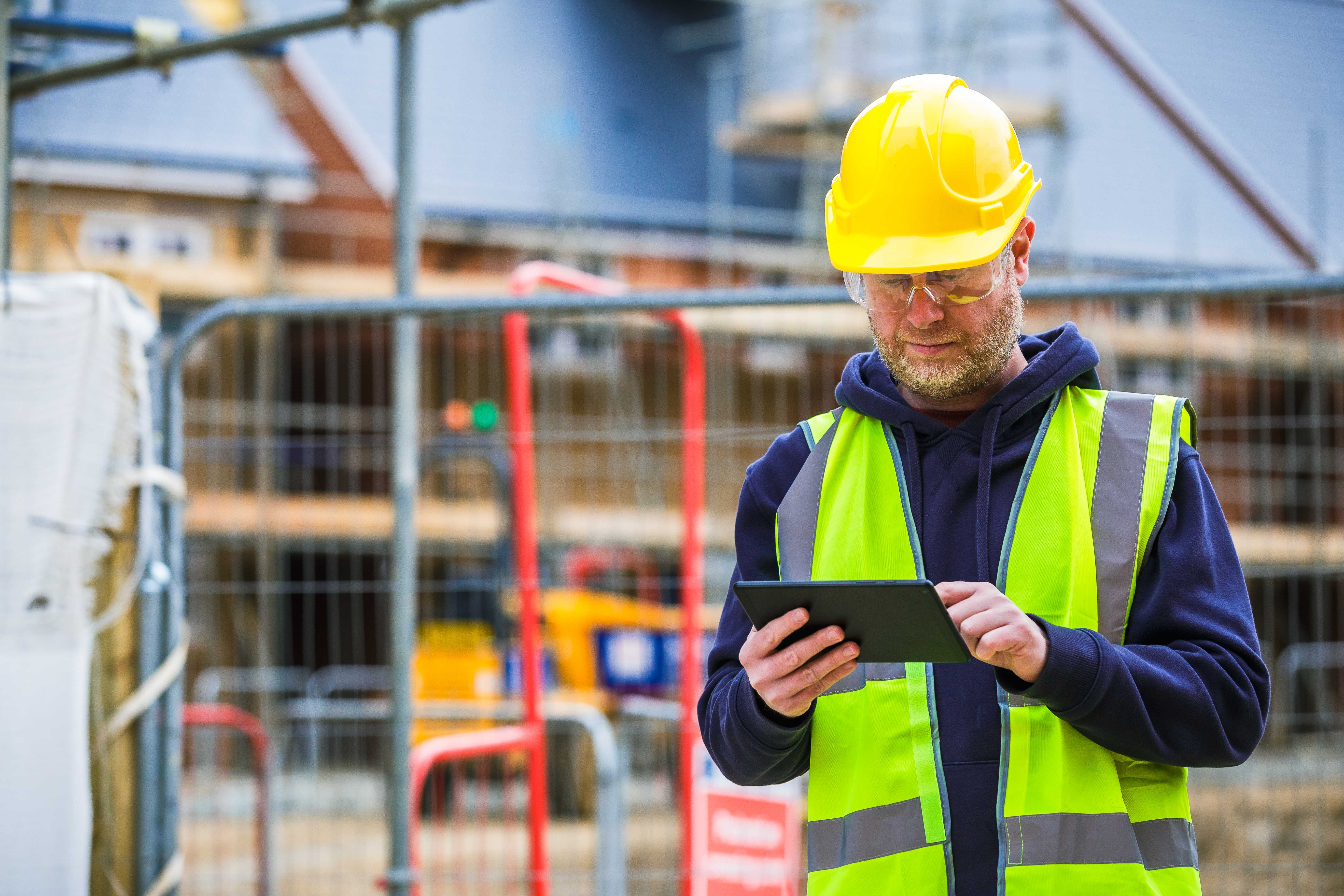 A mid adult male construction worker in full protective workwear - hi visibility vest, goggles and hardhat - using a digital tablet on a construction site where new houses are being built.