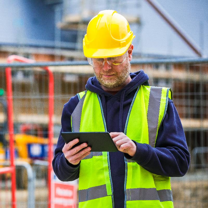 A mid adult male construction worker in full protective workwear - hi visibility vest, goggles and hardhat - using a digital tablet on a construction site where new houses are being built.