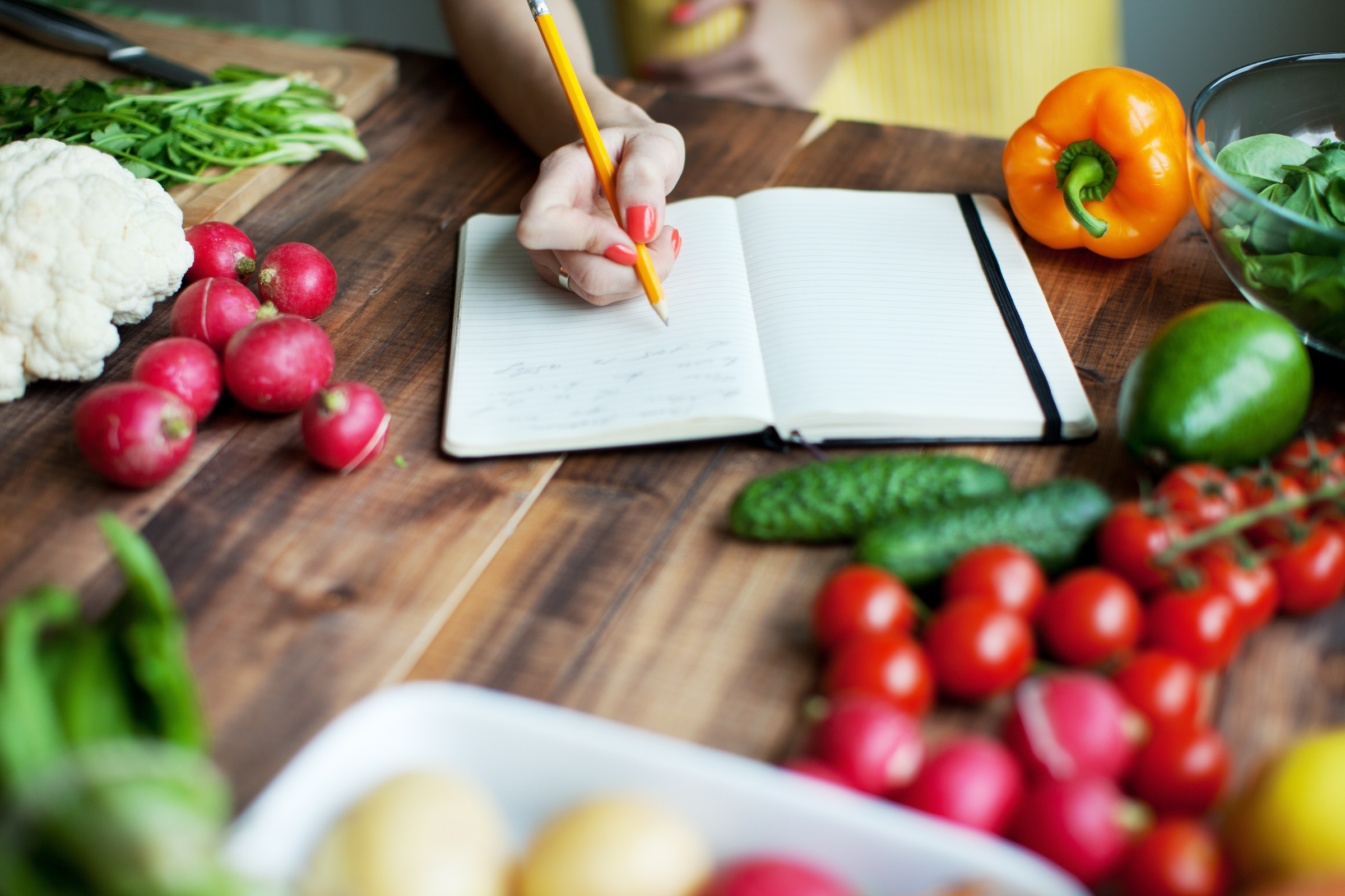 someone writing into a notebook, surrounded by fruit and vegetables