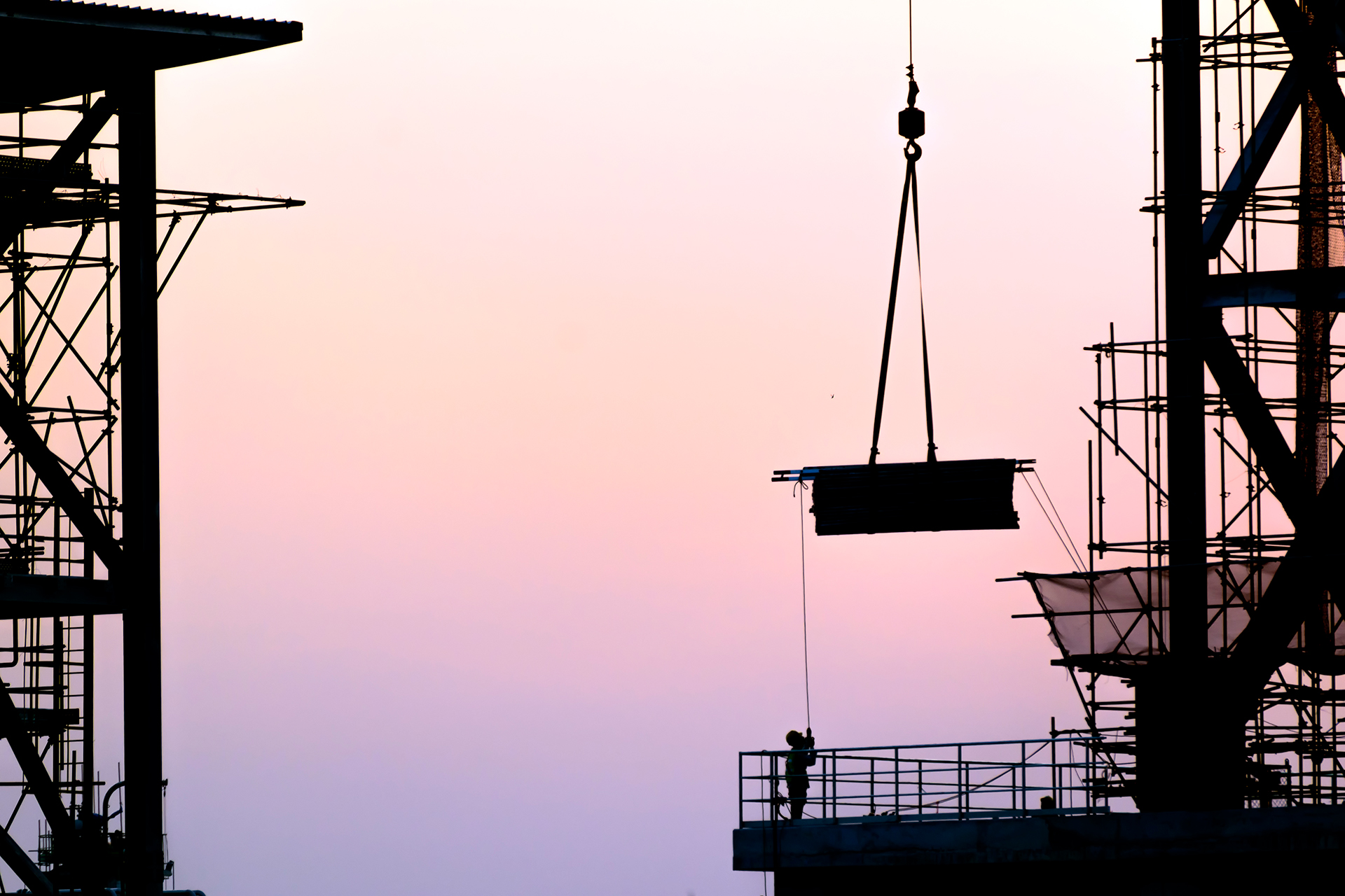 A construction site at dusk with a crane lifting a large rectangular object. The silhouette of the crane and scaffolding structures are visible against the gradient sky, which transitions from light pink to purple. A worker stands on a platform, overseeing the operation.