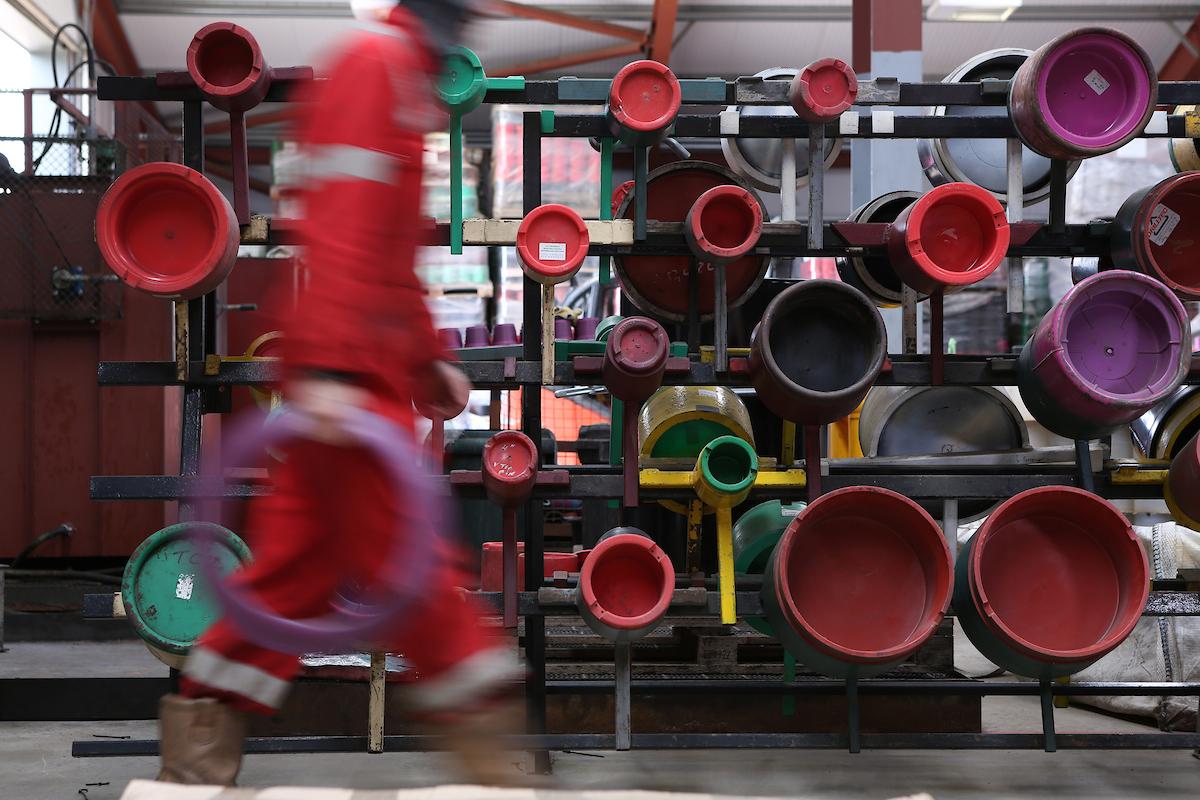 Worker in red safety gear passes blurred in front of a rack filled with stacked pipes and fittings in an industrial warehouse.