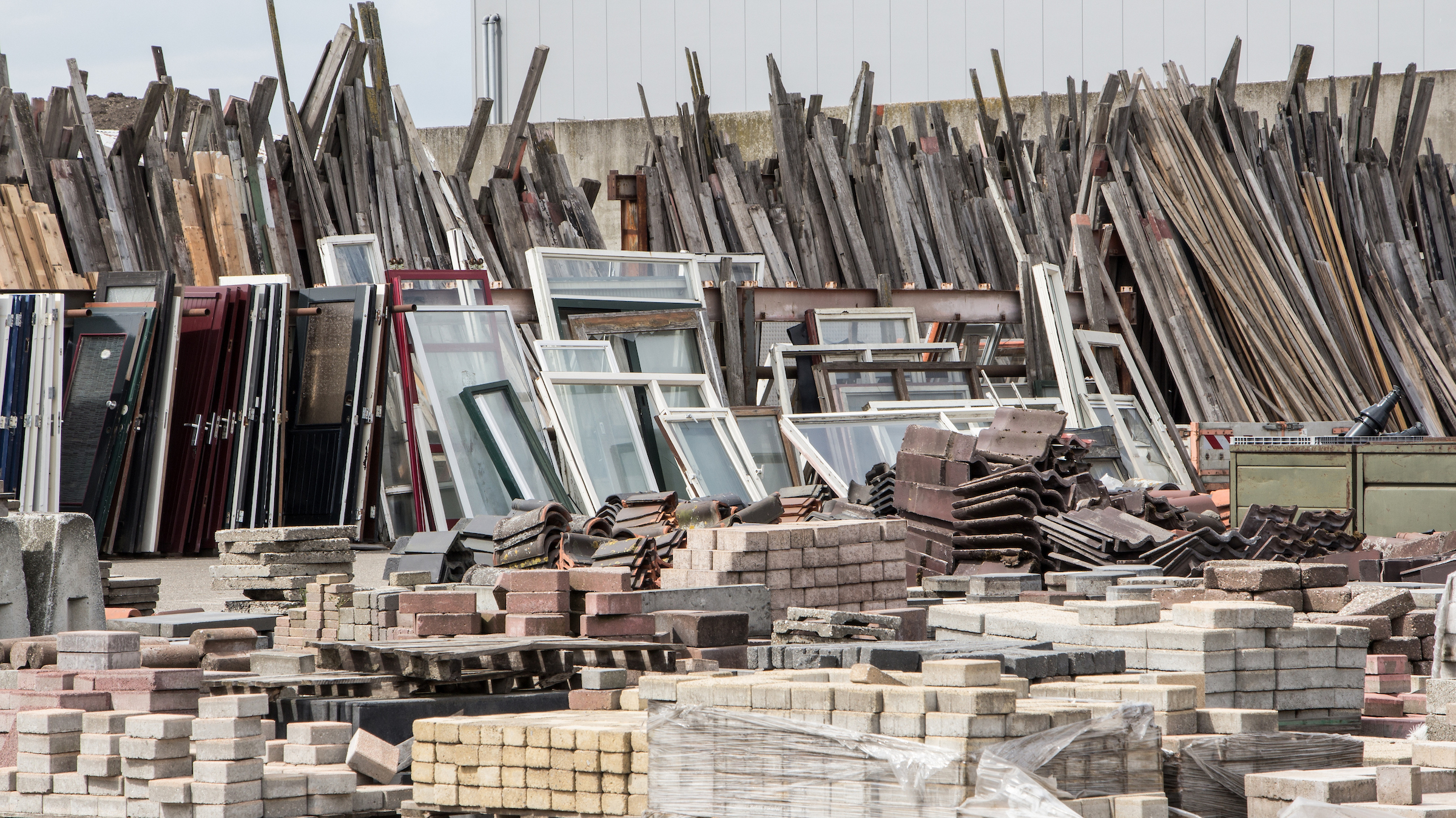 Construction salvage yard with stacked reclaimed timber, window frames, paving blocks, and roofing materials stored outdoors.